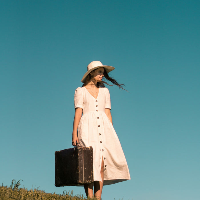 woman standing on green grass holding brown leather suitcase wearing white button-up dress