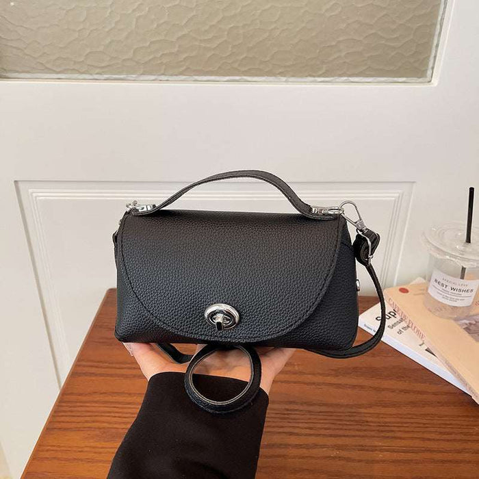 A black pebbled leather handbag with a top handle and shoulder strap, held by a hand over a wooden table in an indoor setting.