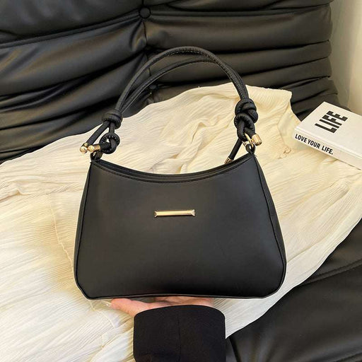 A black leather handbag with knotted top handles and a small gold bar detail on the front, held by a hand over a cream-colored textured fabric with a black leather sofa in the background.