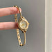 A gold-tone NAOJI watch with a curved bangle bracelet, held by a person's fingers against a neutral gray background.