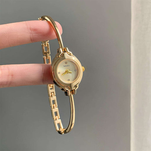 A gold-tone NAOJI watch with a curved bangle bracelet, held by a person's fingers against a neutral gray background.