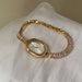 A gold-tone women's watch with a bezel-encased oval face and a link bracelet covered in clear rectangular crystals, resting on a white textured fabric surface.