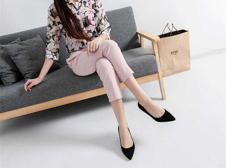Woman sitting on a gray sofa wearing pink pants and black shoes, with a beige bag beside her.