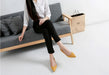 Woman sitting on a gray sofa wearing black pants and yellow shoes, with a beige bag beside her.