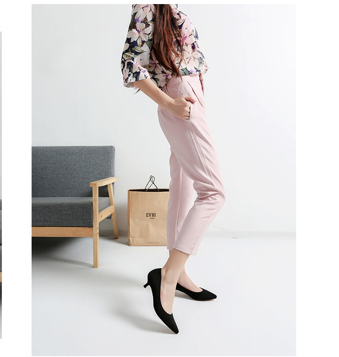 Woman in pink pants and floral top standing in a minimalistic room with a gray chair and brown bag.