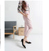 Woman in pink pants and floral top standing in a minimalistic room with a gray chair and brown bag.