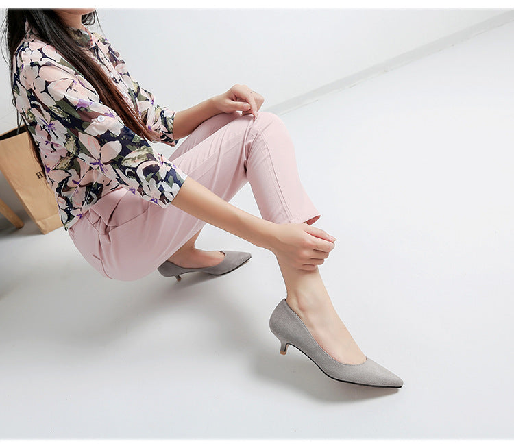 Woman sitting on a white floor wearing floral blouse, pink pants, and gray high heels.