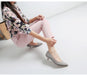 Woman sitting on a white floor wearing floral blouse, pink pants, and gray high heels.