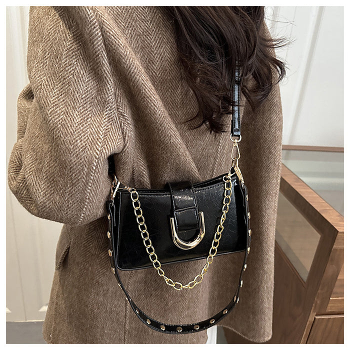 A black faux-leather shoulder bag with a gold chain strap and silver-tone studs, worn crossbody by a person wearing a brown herringbone coat.