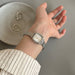 A silver-toned women's watch with a square case and mesh metal band, worn on a person's wrist. The watch has a white dial with gold-tone hour markers.
