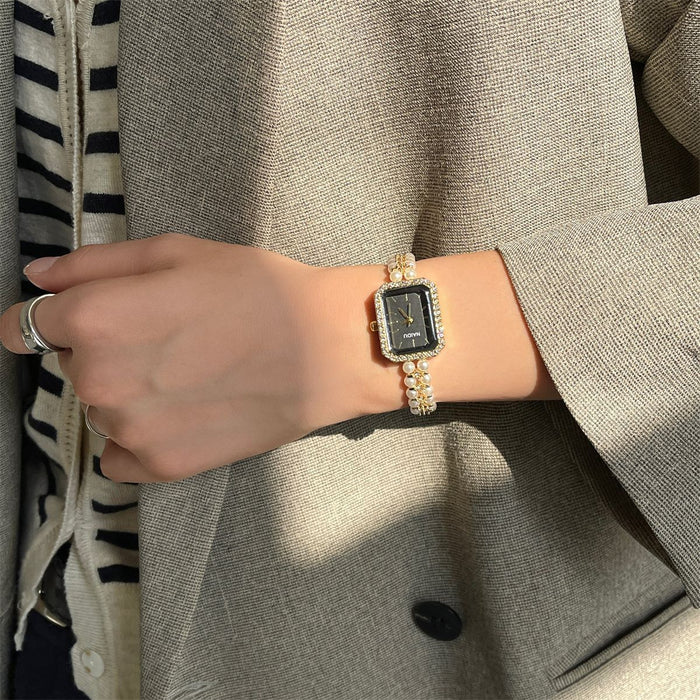 A close-up of a person's wrist wearing a rectangular watch with a black dial and a gold-tone bezel and band embellished with small clear stones, styled with a beige blazer.