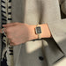 A close-up of a person's wrist wearing a rectangular watch with a black dial and a gold-tone bezel and band embellished with small clear stones, styled with a beige blazer.