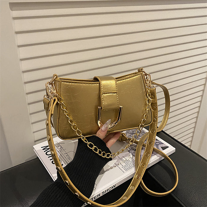 A metallic gold shoulder bag with a chain strap and a studded strap detail, held by a hand over a magazine on a black surface in front of a white slatted wall.