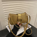A metallic gold shoulder bag with a chain strap and a studded strap detail, held by a hand over a magazine on a black surface in front of a white slatted wall.