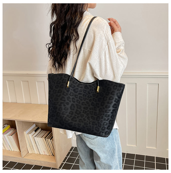 A woman wearing a cream-colored sweater and light blue jeans carries a large black leopard-print tote bag with gold-tone hardware over her shoulder in an indoor setting.
