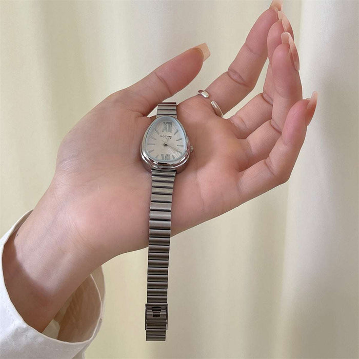 A silver-toned women's wristwatch with an oval silver-tone metal link bracelet, held in a person's hand against a soft cream-colored curtain background.