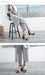 Woman sitting on a stool wearing white pants and black high-heeled sandals in a minimalistic setting.