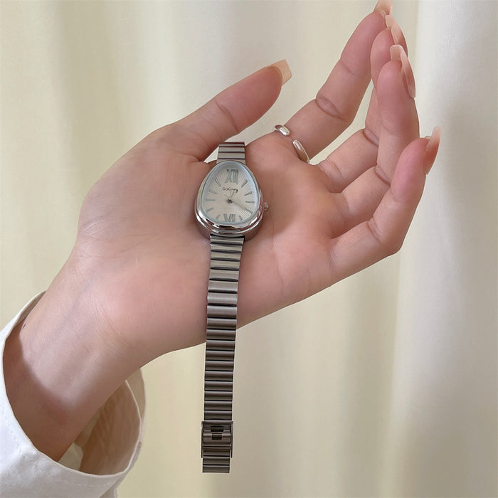 A silver-toned women's wristwatch with an oval silver-tone metal link bracelet, held in a person's hand against a soft cream-colored curtain background.