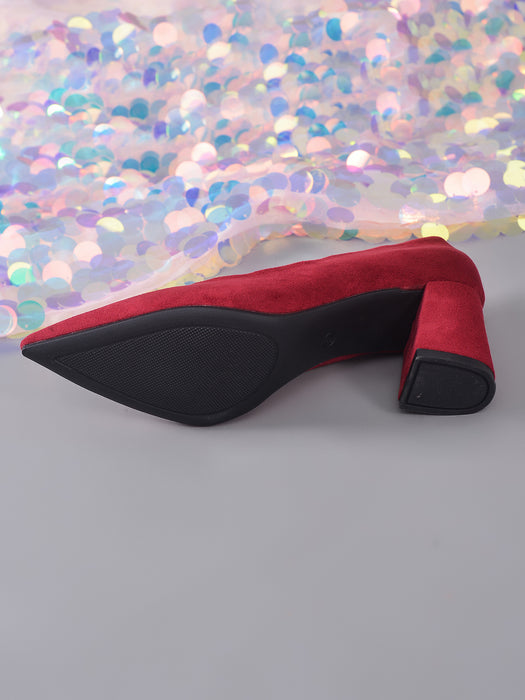 A red suede pointed-toe high heel shown from the side, with a black sole and block heel, placed on a gray surface in front of a shimmering iridescent fabric backdrop.