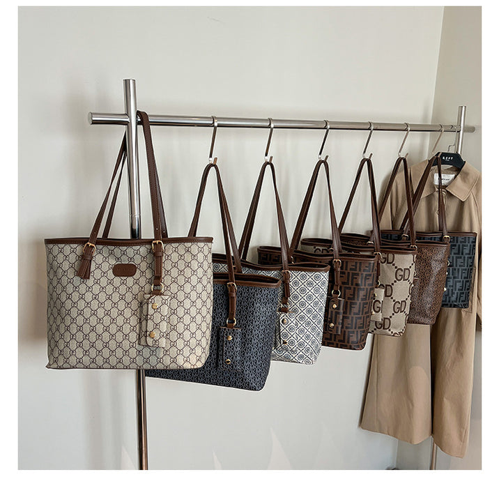 Several designer tote bags with brown leather straps hang from a metal display rack against a plain light wall. The bags feature monogram patterns in beige, navy, gray, and brown, with a beige trench coat hanging on the right.