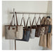 Several designer tote bags with brown leather straps hang from a metal display rack against a plain light wall. The bags feature monogram patterns in beige, navy, gray, and brown, with a beige trench coat hanging on the right.