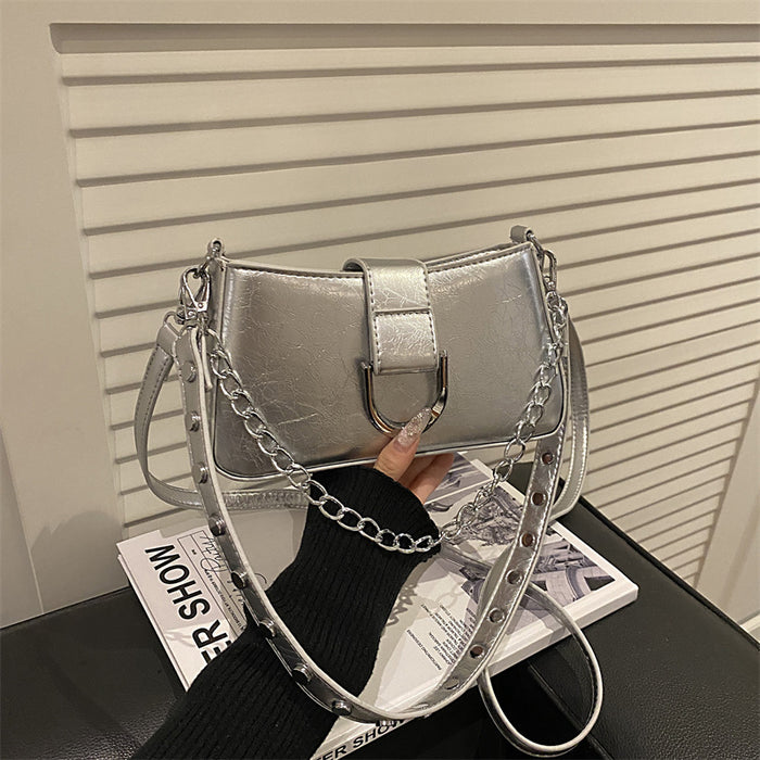 A metallic silver shoulder bag with a chain strap and studded details, held by a hand wearing a black sweater in front of a white slatted wall.