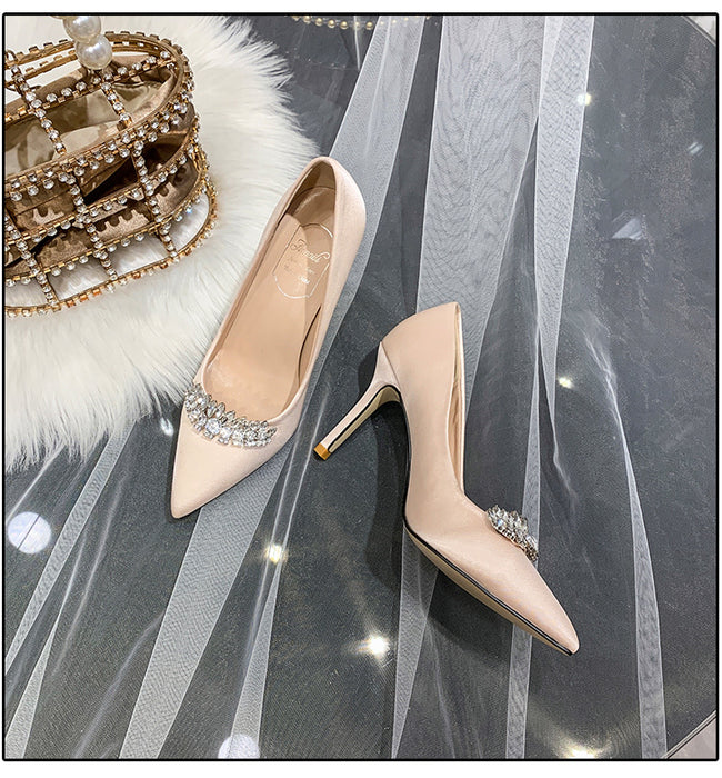Beige high-heeled shoes with crystal embellishments on a gray surface with a decorative bag.