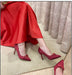 Red high-heeled shoes with decorative elements worn by a person sitting on a reflective surface.