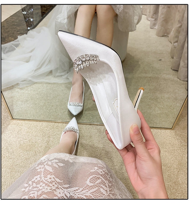 White high-heeled shoes with embellishments held by a hand, worn by a person in a white dress.