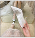 White high-heeled shoes with embellishments held by a hand, worn by a person in a white dress.