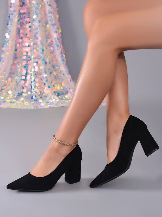 Black pointed-toe block-heel pumps with an ankle chain detail, shown on a model's feet against a gray studio background with a sequined dress prop.