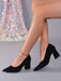 Black pointed-toe block-heel pumps with an ankle chain detail, shown on a model's feet against a gray studio background with a sequined dress prop.