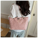 A woman wearing a white blouse and light blue jeans carries a pink tote bag with a repeating black monogram pattern and a gold chain strap in an indoor setting.