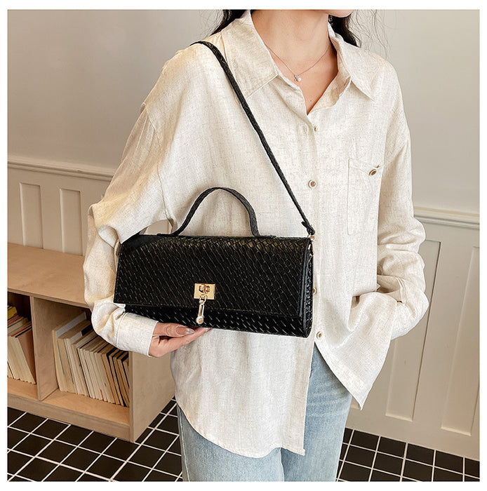 A person wearing a light beige linen shirt and light blue jeans holds a black textured shoulder bag with a gold clasp. The setting is a home interior with a light wood bookshelf and paneled wall.