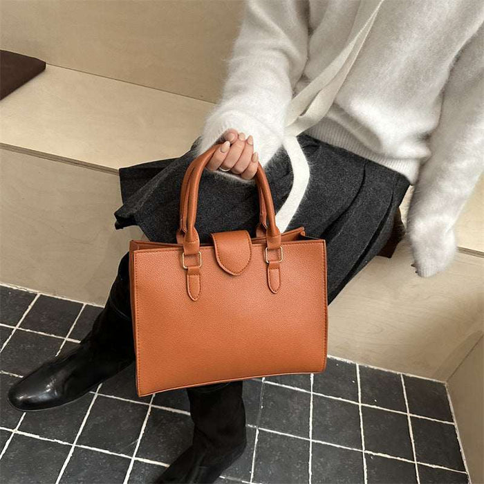 A person in a white sweater and dark skirt holds a structured tan leather tote bag with double top handles and buckle details, seated on a bench in a modern interior with black-and-white tiled flooring.
