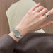 A close-up of a woman's hand wearing a silver-toned metal link watch with a gray dial and a silver signet ring with a black oval stone, set against a soft beige and brown background.