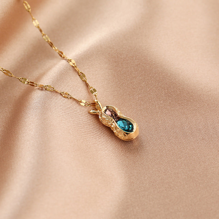 A gold chain necklace with a pear-shaped pendant featuring a sparkling blue gemstone and a small rose-gold accent, displayed on a soft beige satin fabric.