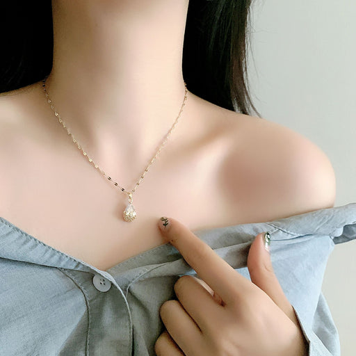 A close-up of a woman wearing a delicate gold chain necklace with a teardrop-shaped clear crystal pendant, styled with a light blue off-the-shoulder shirt.