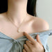 A close-up of a woman wearing a delicate gold chain necklace with a teardrop-shaped clear crystal pendant, styled with a light blue off-the-shoulder shirt.