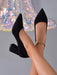 A pair of black pointed-toe high heels with block heels, shown on a model's feet against a gray background with a sparkly iridescent fabric prop.