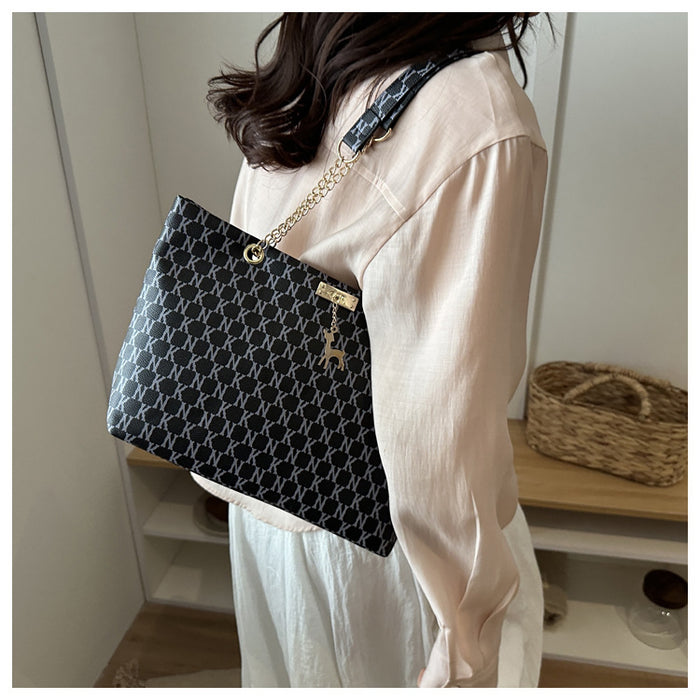 A person wearing a cream-colored blouse and white skirt carries a black shoulder bag with a white geometric monogram pattern, a gold chain strap, and a small gold hanging charm in an indoor setting.