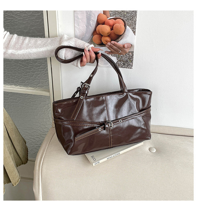 A dark brown faux-leather handbag with dual top handles and multiple zippered compartments, held by a person in a cozy indoor setting with a beige armchair and a framed picture in the background.