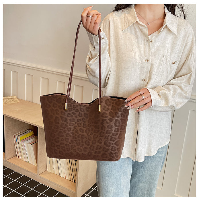 A person wearing a cream button-up shirt and light blue jeans holds a brown leopard-print tote bag with dark brown shoulder straps in an indoor room setting.
