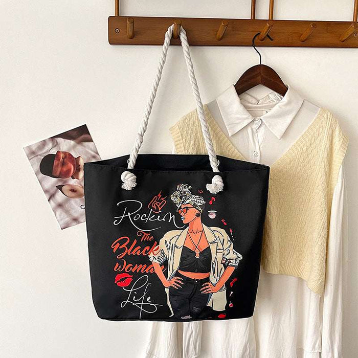 Black tote bag with a graphic design of a woman and text, hanging on a wooden coat rack with a shirt and photo in the background.