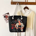 Black tote bag with a graphic design of a woman and text, hanging on a wooden coat rack with a shirt and photo in the background.