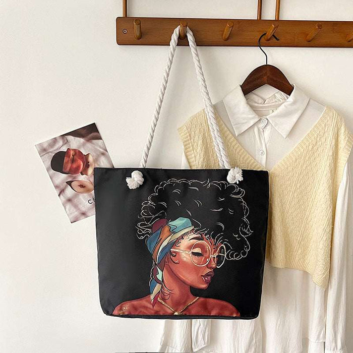 Black tote bag with a colorful design of a woman's face, hanging on a wooden hook with a white shirt and photo album in the background.