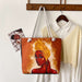 Tote bag with a painted design of a woman with a yellow headscarf, hanging on a wooden rack with other items.