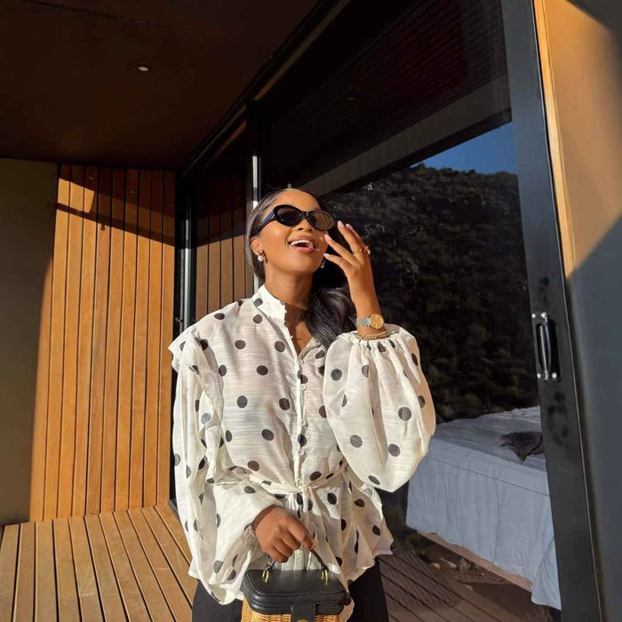 A woman wearing a white long-sleeve blouse with dark gray polka dots, black sunglasses, and a gold watch, standing on a wooden deck in front of a modern house with a view of a mountain landscape.