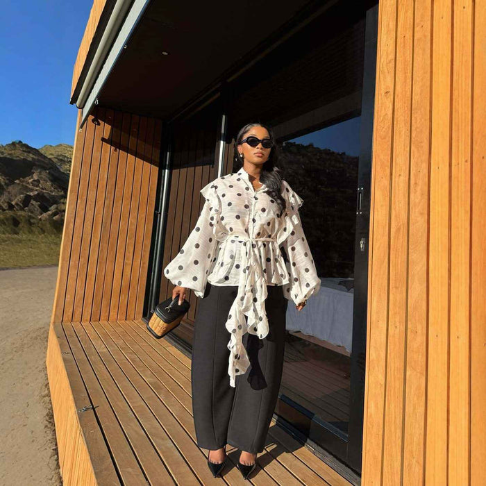 A woman stands on a wooden deck in front of a modern wood-paneled building, wearing a white long-sleeve blouse with black polka dots and a tie waist, paired with dark wide-leg pants and black heels.