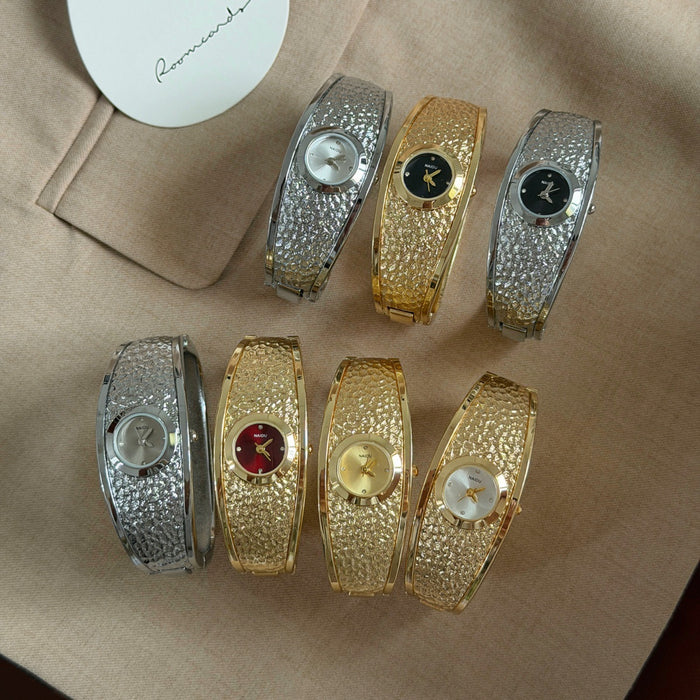 Six textured bangle-style watches arranged on a beige fabric surface, with two silver-toned and two gold-toned bracelets plus two additional silver and gold bracelets showing different dial colors. A white circular card with the brand name Reminisce is partially visible in the top left.